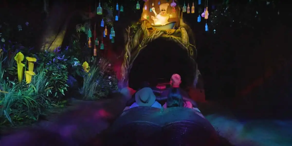 A group of people on Tiana's Bayou Adventure enter a dark tunnel surrounded by lush, colorful foliage. Ambient lighting adds a magical atmosphere, while a prominent glowing figure above the tunnel appears to be a witch or sorcerer casting a spell.